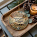 A wooden tray with a decorative ornament featuring a scenic image of Fremont Indian State Park and Museum in Utah, accompanied by pine cones and a lit candle.