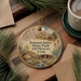 A circular ornament with a scenic illustration of Fremont Indian State Park and Museum, Utah, is displayed on a wooden surface alongside a cup of coffee and some pine branches.