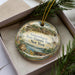 A decorative ornament in a box, featuring a scenic image of the Ganondagan State Historic Site in New York.