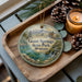 A wooden tray with a decorative ornament featuring a scenic landscape of Giant Springs State Park, Montana, and pine cones.