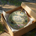 A decorative ornament in a box, featuring a scenic landscape with a river, trees, and wildlife, including a bird and deer.