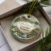 A decorative ornament in a box, featuring a scenic landscape with mountains, water, and flowers.