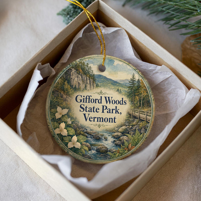 A round ornament with a scenic landscape of a mountain, river, and flowers, displayed in a box with a white tissue paper lining.