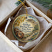 A round ornament with a picture of a log and a path, placed in a box with a white tissue paper lining.