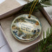 A decorative ornament in a box, featuring a scenic landscape with a lake, mountains, and a bird.