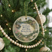 A round ornament hanging on a Christmas tree, featuring a scenic landscape with a bird and flowers.