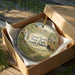 A circular ornament in a wooden box, featuring a scenic landscape with a waterfall, barn, and flowers, with the text "Good Earth State Park, South Dakota" displayed on it.