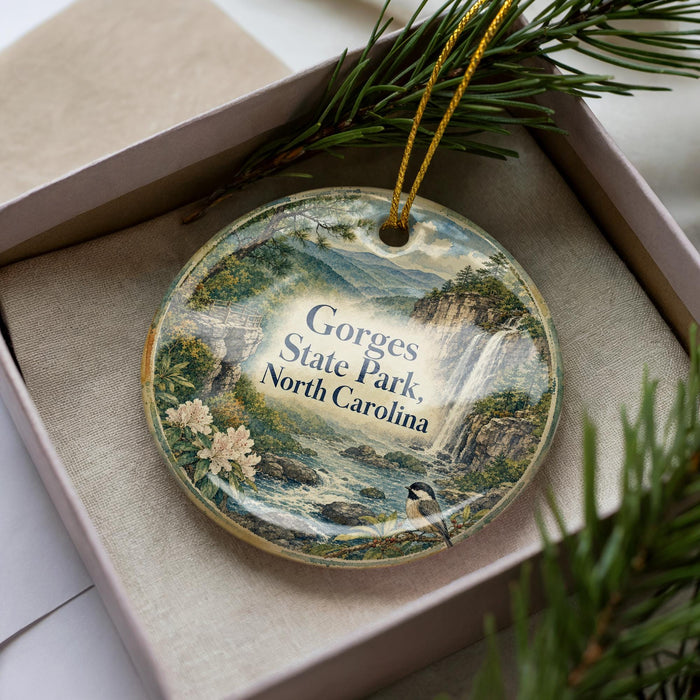 A decorative ornament in a box, featuring a scenic landscape with mountains, waterfalls, and pine trees.