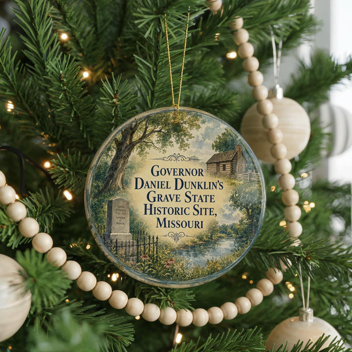 A round ornament hanging on a Christmas tree, featuring a scene of Daniel Dunlin's grave surrounded by trees and a house.