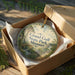 A decorative ornament in a wooden box, featuring a scenic landscape with a bridge and greenery.