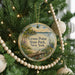 A round ornament hanging on a Christmas tree, featuring a scenic landscape with a lake, trees, and a bird.