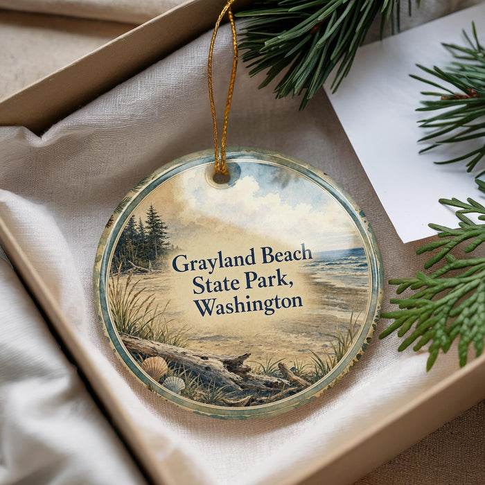 A circular ornament with a painting of a beach scene, featuring a pine tree, grass, and a body of water, is displayed in a box with pine branches.