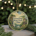 A circular ornament with a scenic landscape featuring a waterfall, a house, and greenery, hanging in front of a blurred background with Christmas lights.