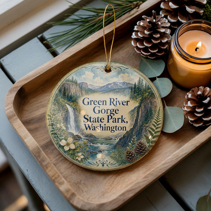 A decorative ornament with a scenic image of the Green River Gorge State Park in Washington, featuring a waterfall and mountains, is displayed on a wooden tray alongside pine cones and a lit candle.