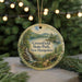 A round ornament with a scenic landscape of a lake, mountains, and a waterfall, surrounded by greenery, hangs in front of a blurred background with twinkling lights.