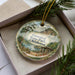 A decorative ornament in a box, featuring a scenic landscape with a lake, mountains, and a cabin.