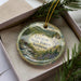 A round ornament with a scenic landscape of the Groton State Forest in Vermont, featuring a river, mountains, and pine trees.