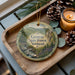 A wooden tray with a decorative ornament featuring a scenic forest landscape, pine cones, and a lit candle.