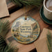 A decorative ornament featuring a scenic image of Guernsey State Park, Wyoming, with a wagon and other natural elements. The ornament is hanging on a wooden surface, surrounded by pine branches and a cup of coffee.