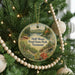 A round ornament hanging on a Christmas tree, featuring a scenic landscape with a lake, forest, and mountains.