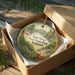 A decorative ornament in a box, featuring a scenic landscape with a lake, forest, and mountains.