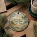 A round ornament with a lighthouse and beach scene, surrounded by pine branches and a cup of coffee on a wooden surface.