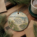 A decorative ornament featuring a beach scene with a lifeguard tower, surrounded by pine branches and flowers, placed on a wooden surface next to a cup of coffee.