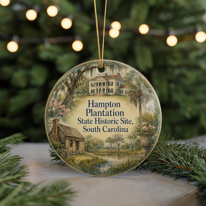 A round ornament with a painting of a historic plantation house and a pond, surrounded by greenery, placed on a surface with Christmas lights in the background.