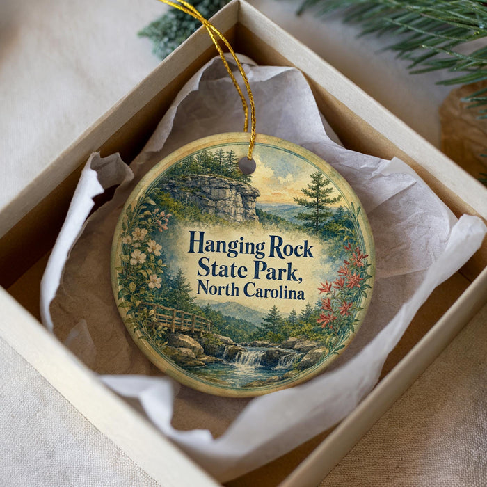 A round ornament with a scenic landscape of hanging rock, state park, and water, displayed in a box with a white tissue paper lining.