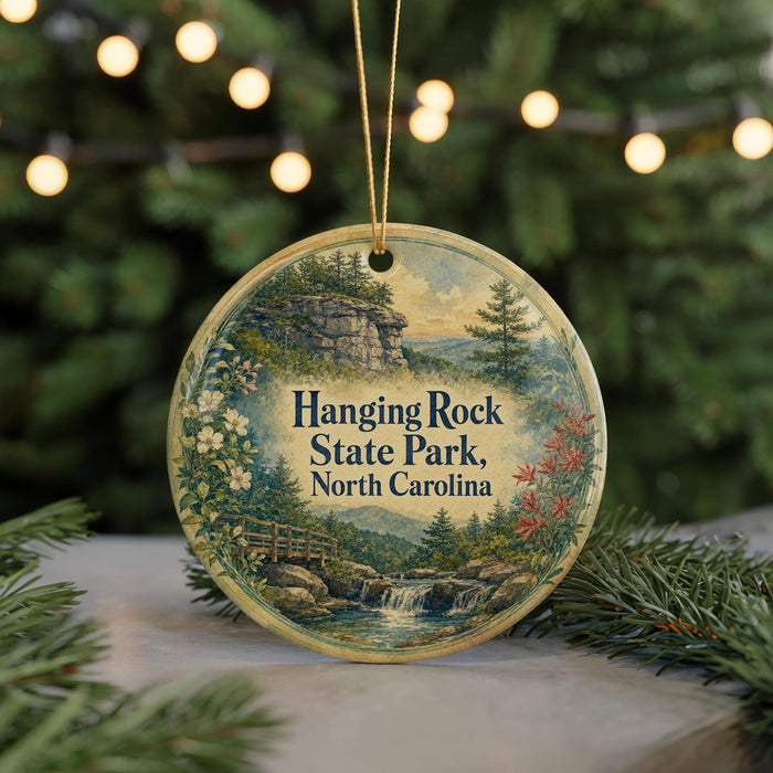 A round ornament with a scenic landscape of hanging rock, state park, and water, surrounded by pine branches and greenery, with a string of lights in the background.