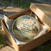 A decorative ornament in a wooden box, featuring a scenic landscape with a lighthouse and trees, with the text "Harrington Beach State Park, Wisconsin" printed on it.