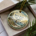 A decorative ornament in a box, featuring a scenic landscape with mountains, water, and pine trees.
