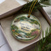 A decorative ornament in a box, featuring a scenic landscape with a river, trees, and a deer.
