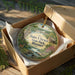 A decorative ornament in a box, featuring a scenic landscape with a river, trees, and a bridge.