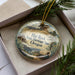 A decorative ornament in a box, featuring a scenic landscape with a cabin and mountains in the background.