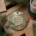 A round ornament with a scenic landscape of a river and mountains, surrounded by pine branches, sits on a wooden surface.