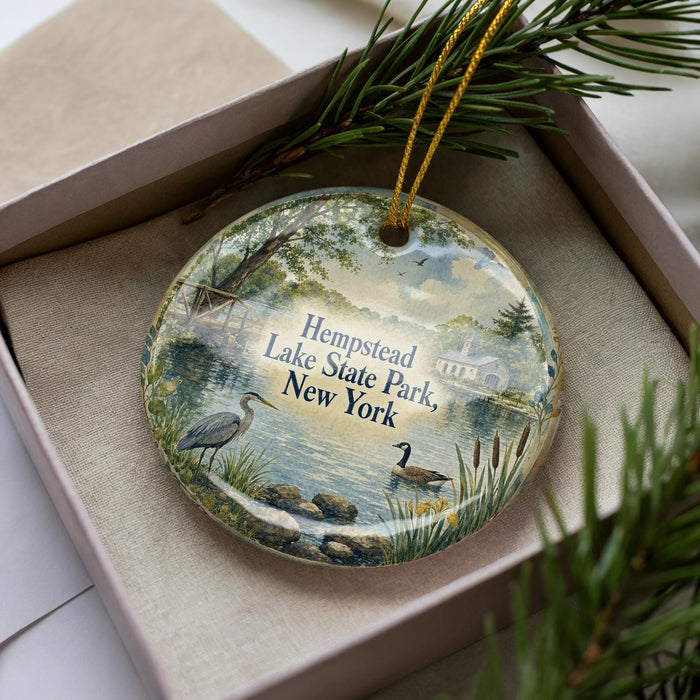 A decorative ornament with a lake and park scene, featuring a bird and a duck, is displayed in a box with pine branches.