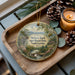A decorative ornament with a scenic landscape of Hickory Knob State Resort Park, South Carolina, is displayed on a wooden tray alongside pine cones and a lit candle.