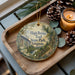 A decorative ornament with a scenic image of the High Bridge Trail in Virginia, featuring a bridge, trees, and a river, is displayed on a wooden tray alongside pine cones and a lit candle.