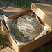 A decorative ornament in a wooden box, featuring a scenic landscape with a river, mountains, and wildlife.
