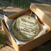 A round ornament in a wooden box, featuring a scenic landscape with trees and a body of water.