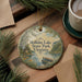 A round ornament with a scenic image of Holliday Lake State Park, Virginia, hangs on a wooden surface. The ornament is surrounded by pine branches and a cup of coffee, creating a cozy and festive atmosphere.