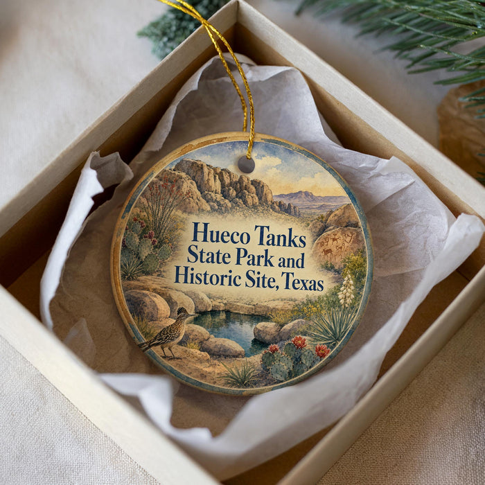 A round ornament with a painting of a bird and a desert landscape, placed in a box with a white tissue paper lining.