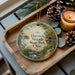 A decorative ornament featuring a scenic image of Hueston Woods State Park, Ohio, with a candle and pine cones on a wooden tray.