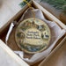 A decorative ornament in a box, featuring a scenic landscape with a waterfall, houses, and greenery, with the text "Icelandic State Park, North Dakota" printed on it.