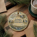 A decorative ornament featuring a scenic landscape of Iceland, including a waterfall and a church, is displayed on a wooden surface alongside a cup of coffee and some pine branches.