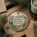 A decorative ornament featuring a scenic landscape with mountains, pine trees, and a lake, hanging on a wooden surface.