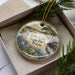 A decorative ornament in a box, featuring a scenic landscape with a lake, trees, and a bird, along with the text "Inks Lake State Park, Texas".