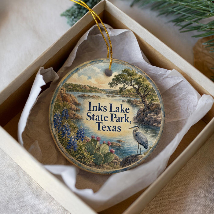 A circular ornament with a painting of a lake and a bird, surrounded by flowers and trees, placed in a box with a white tissue paper lining.
