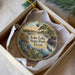 A circular ornament with a painting of a lake and a bird, surrounded by flowers and trees, placed in a box with a white tissue paper lining.